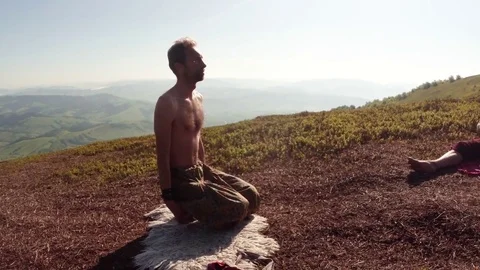 Young guy raises his hands up while sitting on the ground in the mountains, yoga Stock Footage 77786619