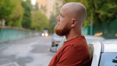 A young guy with a red beard stands in the street leaning against the car Stock Footage 139002293