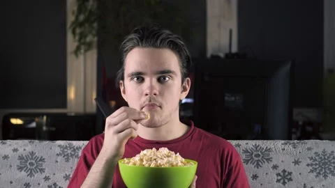 A young guy in a red T-shirt sits on the couch and watches TV, eats popcorn Stock Footage 149627289