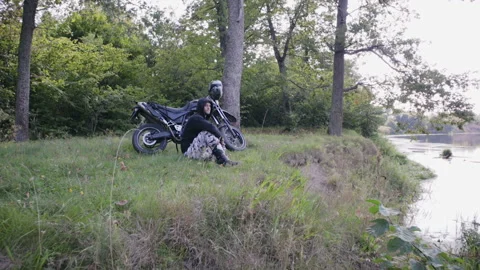 A young guy sits and rests next to his motorcycle on a cliff near the river 動画素材 116228288
