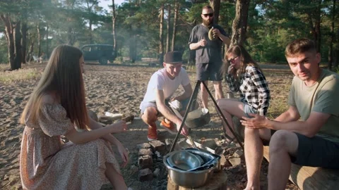 A young guy sits in a circle with his friends and puts firewood on a fire for Stockbeeldmateriaal 237148490