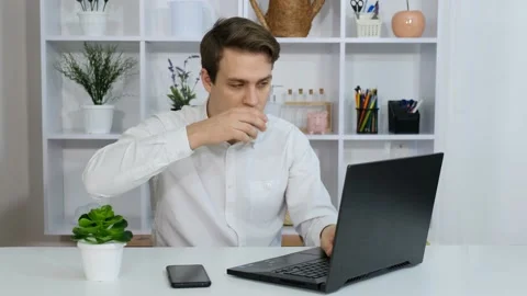 A young guy sits at his desk in the office drinking coffee and working Stock Footage 172392087