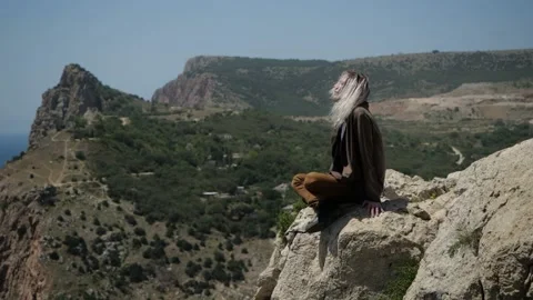 A young guy sits on a mountainside against a panorama of mountains Stock Footage 147908054