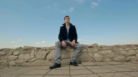 A young guy sits on the stone freely in a black coat Video stock 90216276