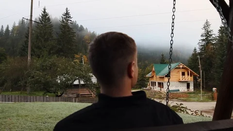 A young guy sits on a swing and looks at the forest in the fog Stock Footage 96223007