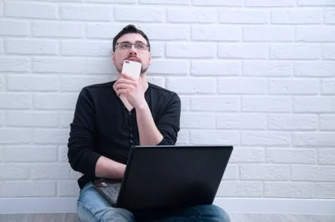 A young guy sits thoughtfully with a computer while holding a phone in his ha Stock Photos
