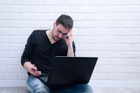 A young guy sits thoughtfully with a computer while holding a phone in his ha Stock Photos