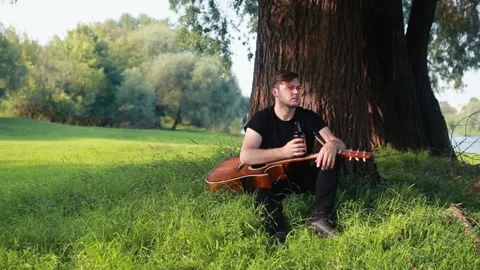 A young guy sits under a tree in his hands holds a guitar and drinks a bottle of Stock Footage 132426306