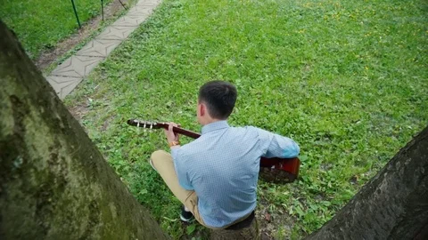 Young guy sitting alone on the grass and playing an acoustic guitar. Stock Footage 70877431