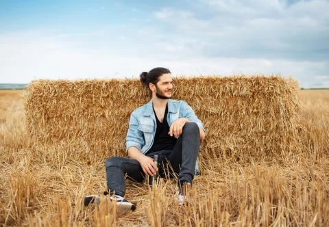Young guy sitting on background of haystacks in wheat field. Stock Photos