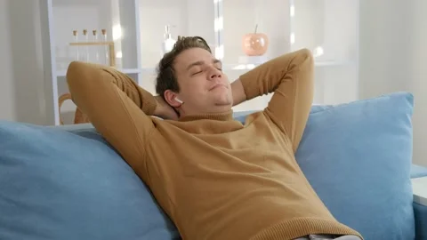 A young guy sitting on the couch inserts headphones into his ears Stock Footage 170864577