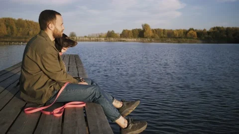 Young guy sitting with dog on the pier and admiring the beauty of the lake Stock Footage 140736033
