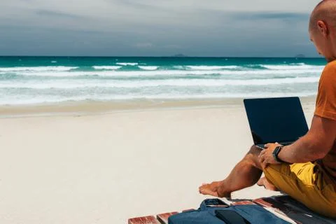 Young guy sitting with his back by the sea on a sunny day, working Stock Photos