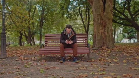 Young guy sitting on a park bench with a phone in his hands.Green tree young man Stock Footage 141808360
