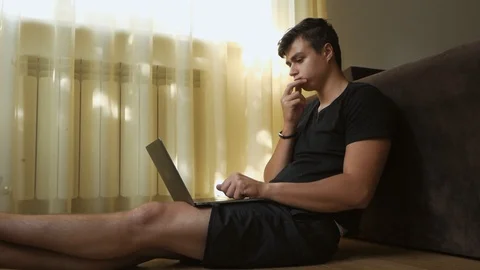 A young guy sitting in a room on the floor by the window uses a laptop. A Stock Footage 108773483