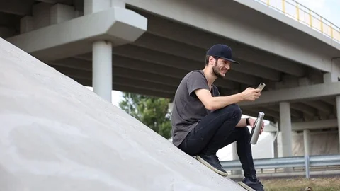 Young guy is sitting under the bridge with smartphone and steel bottle I hands. Stock Footage 115737093