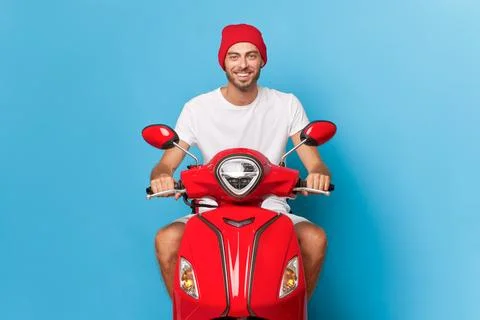 Young guy with small beard in white t-shirt and red hat rides red scooter Stock Photos