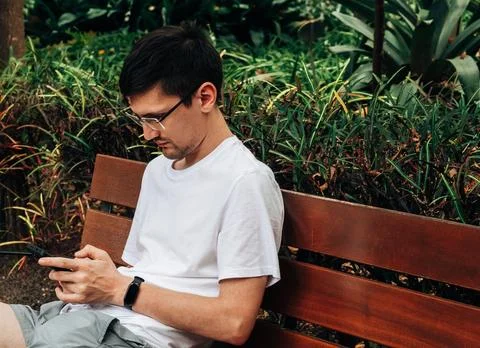 A young guy with a smartphone is sitting on a bench in the garden. Stock Photos