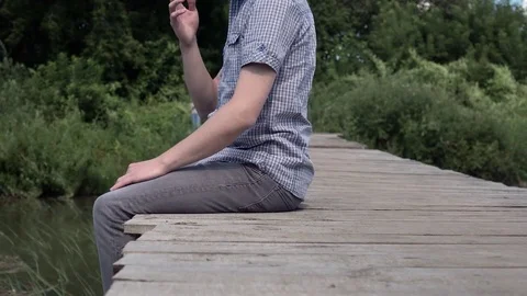 Young guy smokes a cigarette while sitting on a bridge enjoys nature close-up 4k Stock Footage 79125176