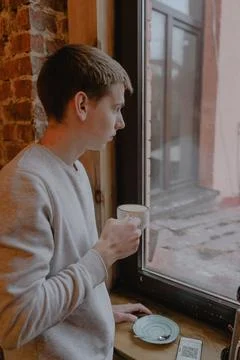 A young guy stands in a cafe by the window with a cup of coffee in his hands Stock Photos