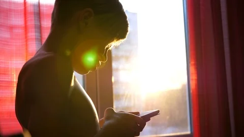 A young guy stands near a window with a phone in his hands. Sunset time, the sun 스톡 동영상 87503864