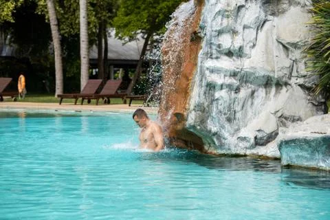 A young guy stands under artificial waterfalls in the pool on site Foto stock