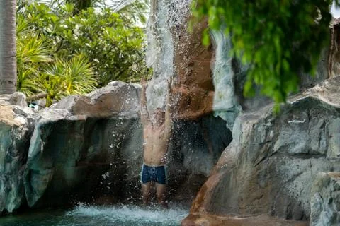 A young guy stands under artificial waterfalls in the pool on site Stock Photos