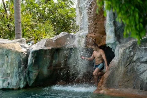 A young guy stands under artificial waterfalls in the pool on site Stock Photos