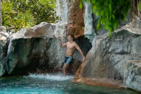 A young guy stands under artificial waterfalls in the pool on site Stock Photos