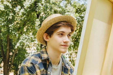 A young guy in a straw hat sits in the park in front of an easel and paints a Stock Photos