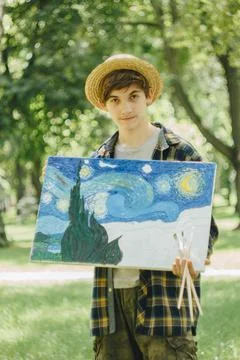 A young guy in a straw hat sits in the park in front of an easel and paints a Stock Photos