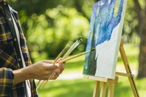 A young guy in a straw hat sits in the park in front of an easel and paints a Stock Photos