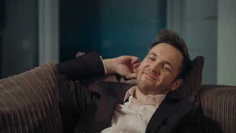 A young guy in a suit sits on a sofa at home and smiles. Stock Footage 219826192