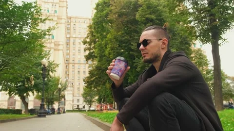 Young guy in sunglasses sits on a border in the park and drinks coffee. Close-up Video stock 139908725