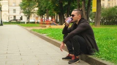Young guy in sunglasses sits on a border in the park and drinks coffee Stock Footage 139910897
