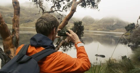 Young guy taking picture on phone of mountain view with lake in foreground Stock Footage 87212289