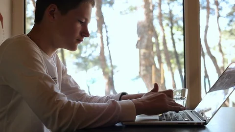 A young guy typing on a laptop in a cafe near the window, a student, drinking wa Stock Footage 114117506