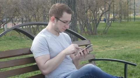 Young guy uses the phone to sit on the bench in the park Video stock 90180681