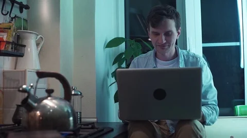 Young guy using computer sitting at kitchen while kettle boiling on stove Stock Footage 83871273