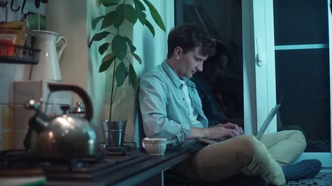 Young guy using computer sitting at kitchen while kettle boiling on stove Video stock 83871517