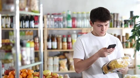 Young guy using smartphone to scan barcode on pasta package in supermarket aisle Stock Footage 345563253