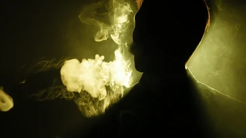 Young guy vaping in the dark room. Silhouette of vaper in the darkness  lighted