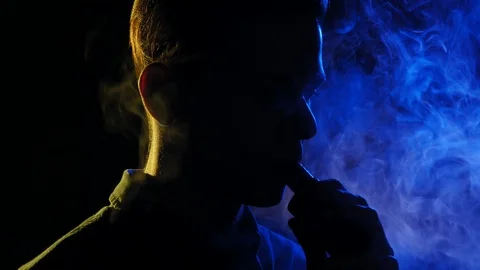 Young guy vaping in the dark room. Silhouette of vaper in the darkness lighted Stock Footage 106471953