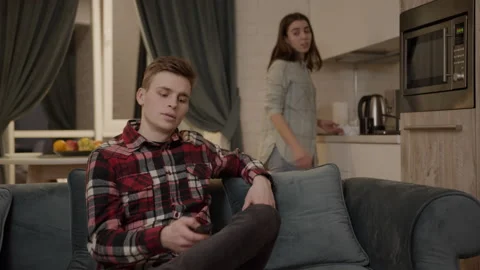 A young guy watches TV while his girlfriend prepares a delicious dinner for them 스톡 동영상 147677694