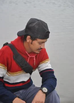 A young guy wearing cap backwards, looking down while sitting next to the lake Stock Photos