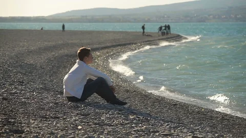 A young guy in a white jacket is sitting on the beach, nervous, puzzled, worried Vidéo 114158015
