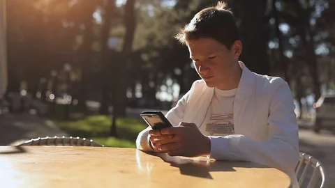 A young guy in white with a smartphone in his hands is sitting at a cafe table o Stock Footage 113672753