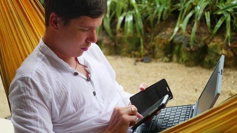 Young guy working on laptop and smartphone lying in a hammock Vidéo 75537953