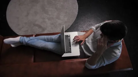 A young guy works remotely on his laptop while lying on the couch. Top Shot Stock Footage 198876808