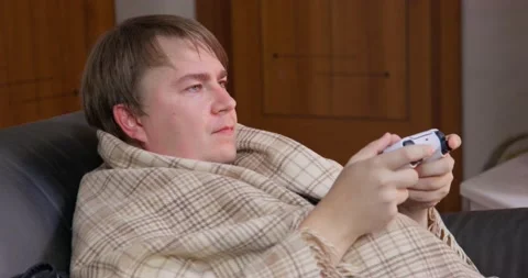 Young guy wrapped in warm blanket is playing a video game holding controller for Video stock 166590533